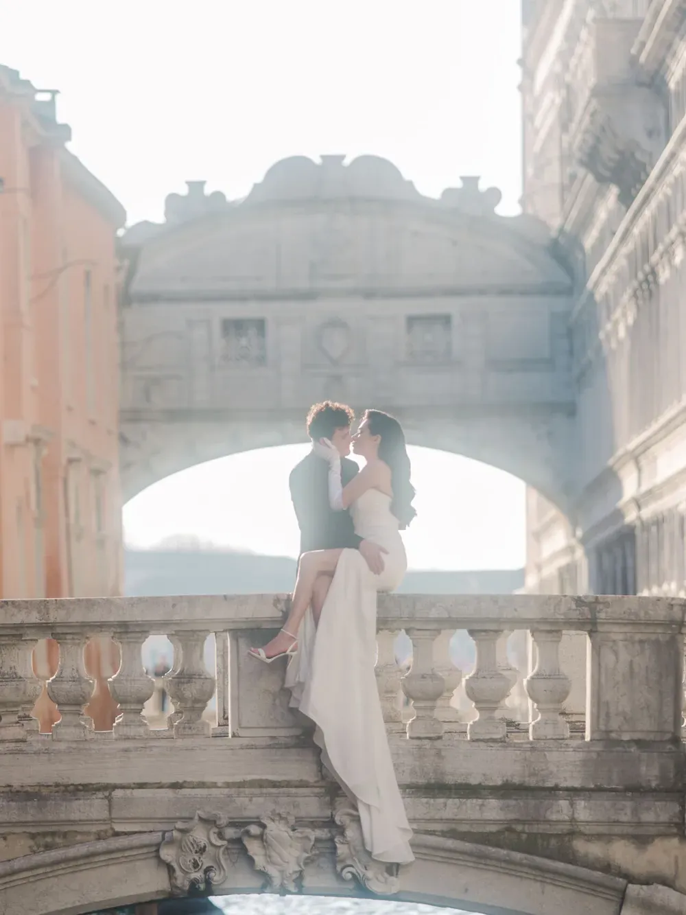 A couple in formal attire sits on a stone balustrade of an ancient Italian bridge, embracing as sunlight reveals the city arches behind them.