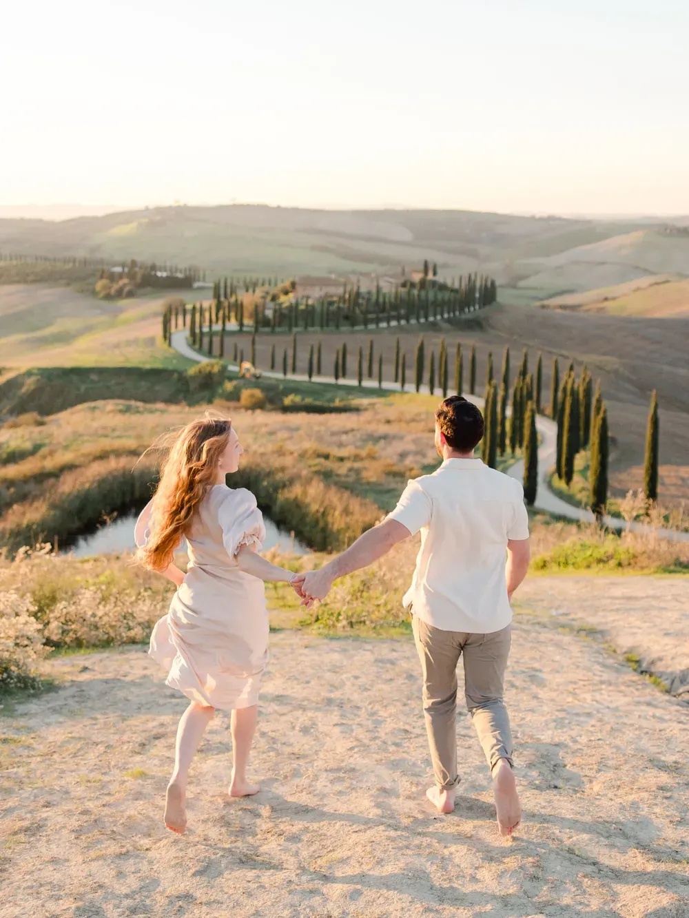 A barefoot couple walks hand-in-hand along a sunlit dirt path through Tuscany’s rolling hills, with a cypress-lined road winding in the distance.