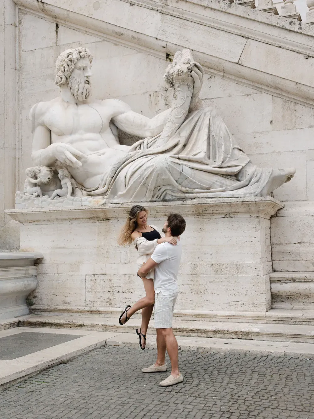 Two people embrace as one lifts the other in front of a large reclining marble statue on a historic Italian plaza.