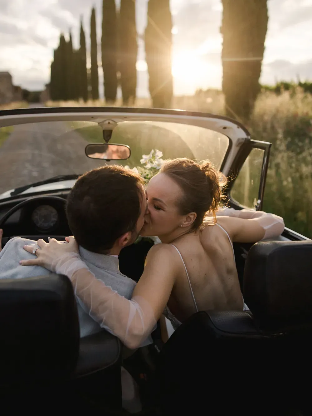 Two people kiss in an open-top car on a sunlit Italian countryside road, with tall cypress trees and a warm sunset.