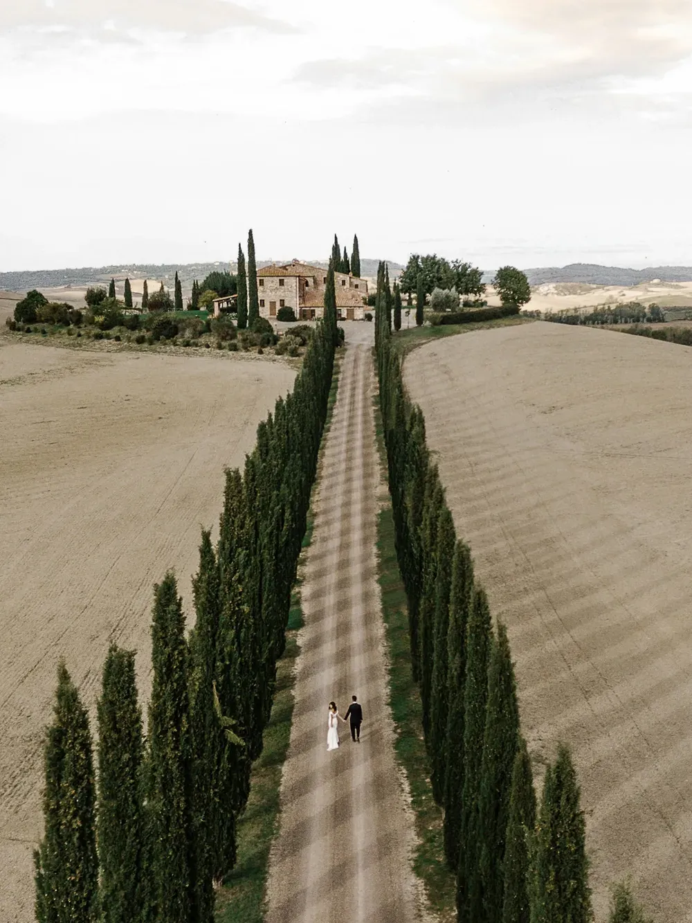 Aerial view of a long gravel road flanked by tall cypress trees leading to a Tuscan farmhouse; a couple in wedding attire walk hand in hand.