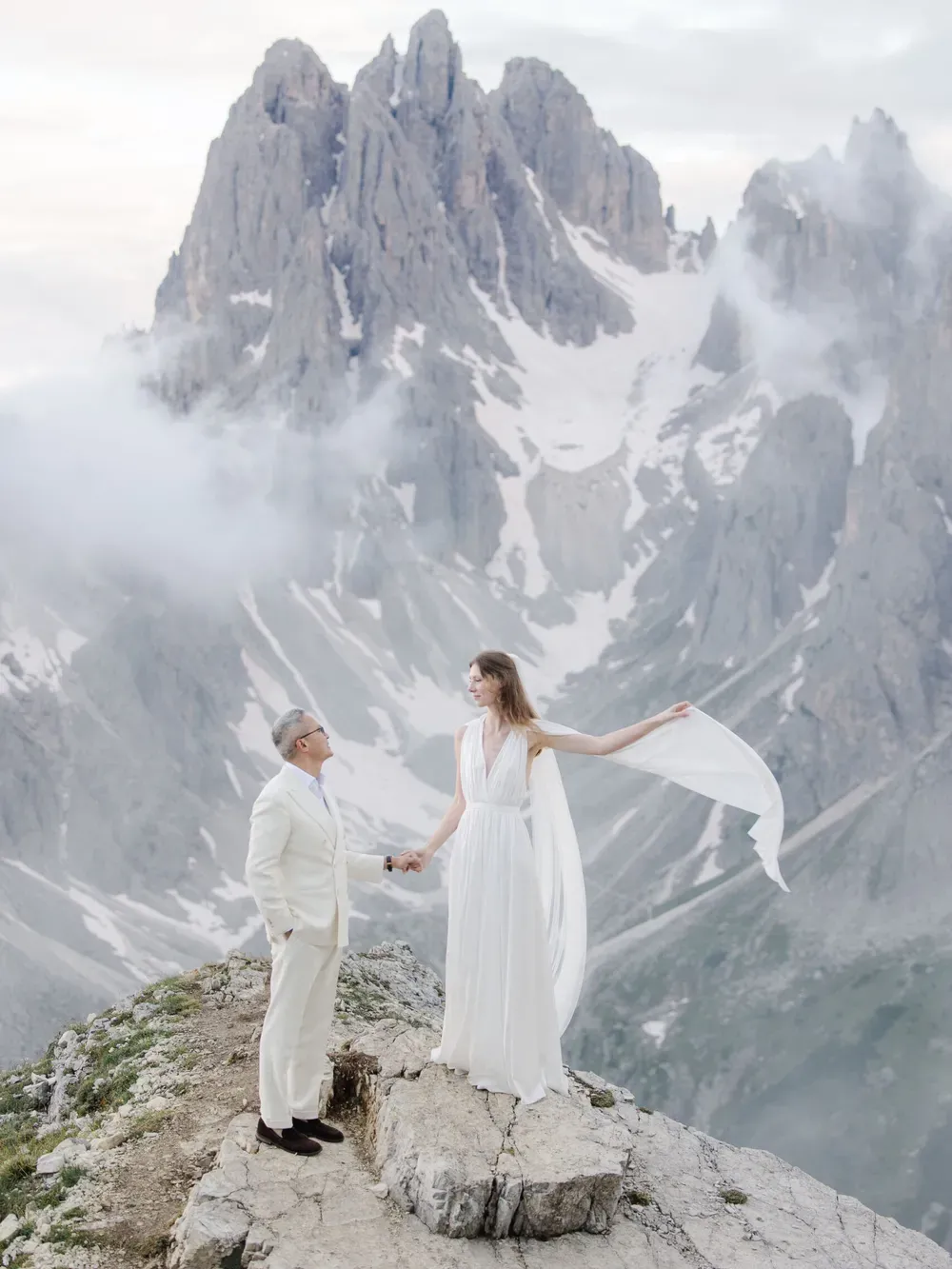 Two people stand on a rocky cliff in white outfits, holding hands as a flowing fabric trails behind, with snow-capped mountains and misty skies.
