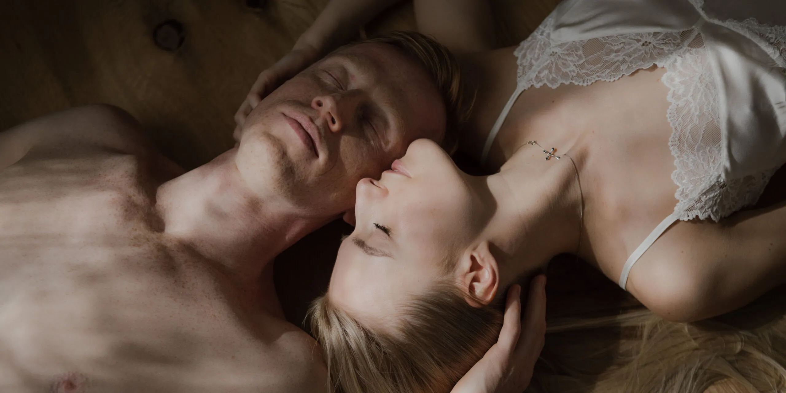 Two people lie on a wooden floor in a tender, intimate pose; one wears white lace lingerie, the other is shirtless, heads close, eyes closed.