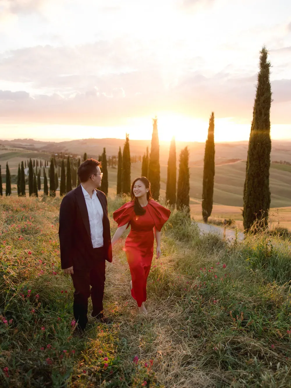 Two people walk hand in hand along a sunlit Tuscan hillside path in Italy, one in a red dress and the other in a dark blazer, with cypress trees at sunset.