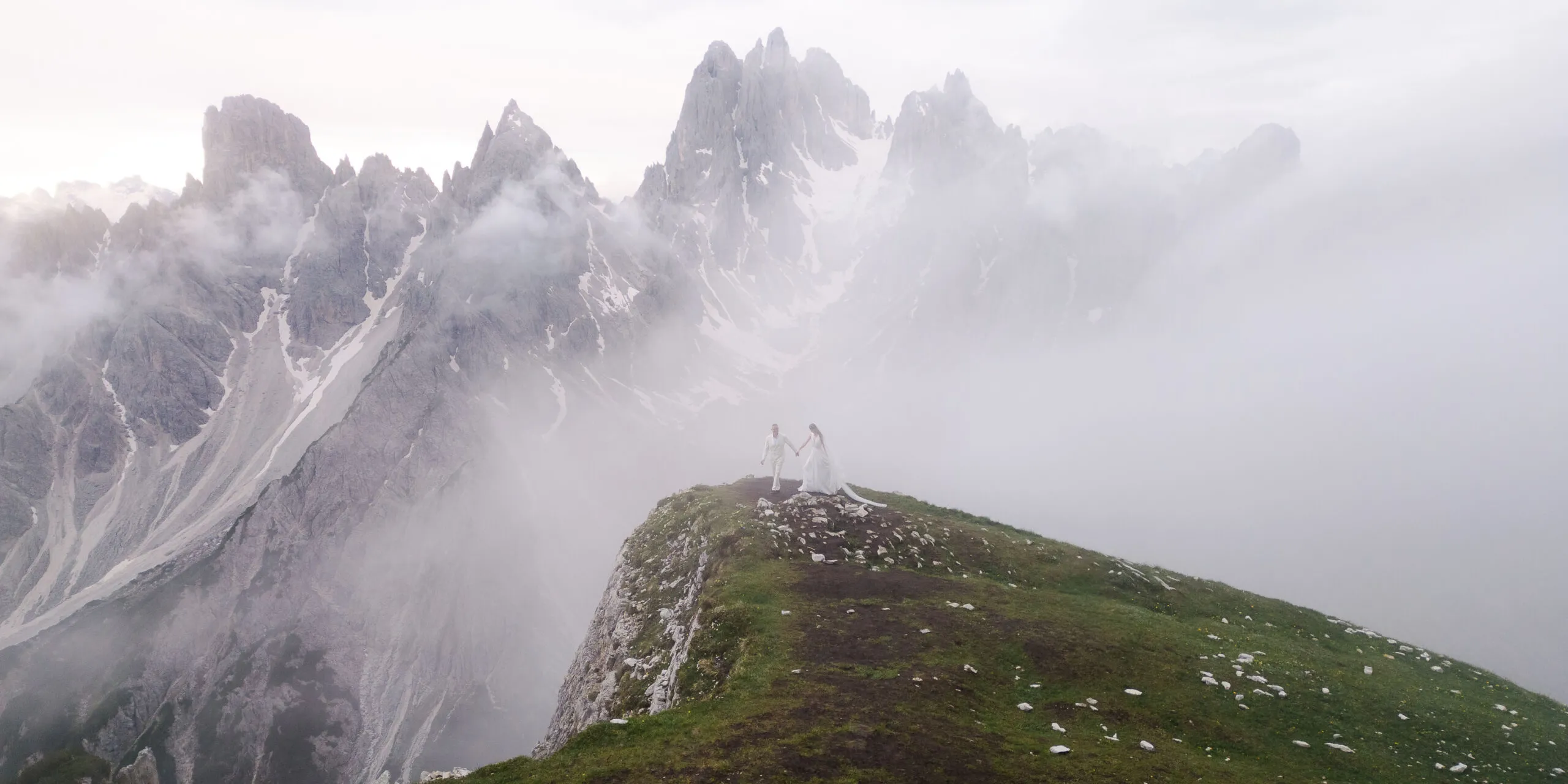 Couple in wedding attire stands on a grassy cliff edge in the Italian Alps, holding hands, with misty peaks rising behind.
