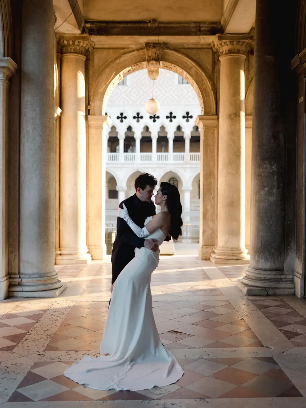 A couple in formal wear share a tender embrace beneath a grand stone arch in a sunlit Italian courtyard; one wears a white wedding gown.