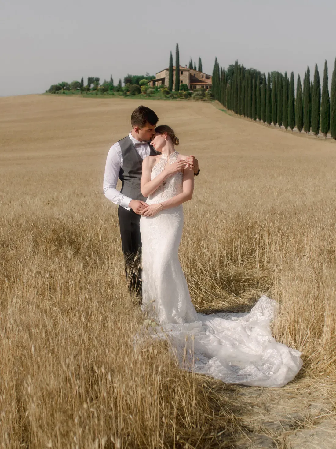 A couple in wedding attire embraces in a golden wheat field, with a Tuscan villa and row of tall cypress trees in the background.