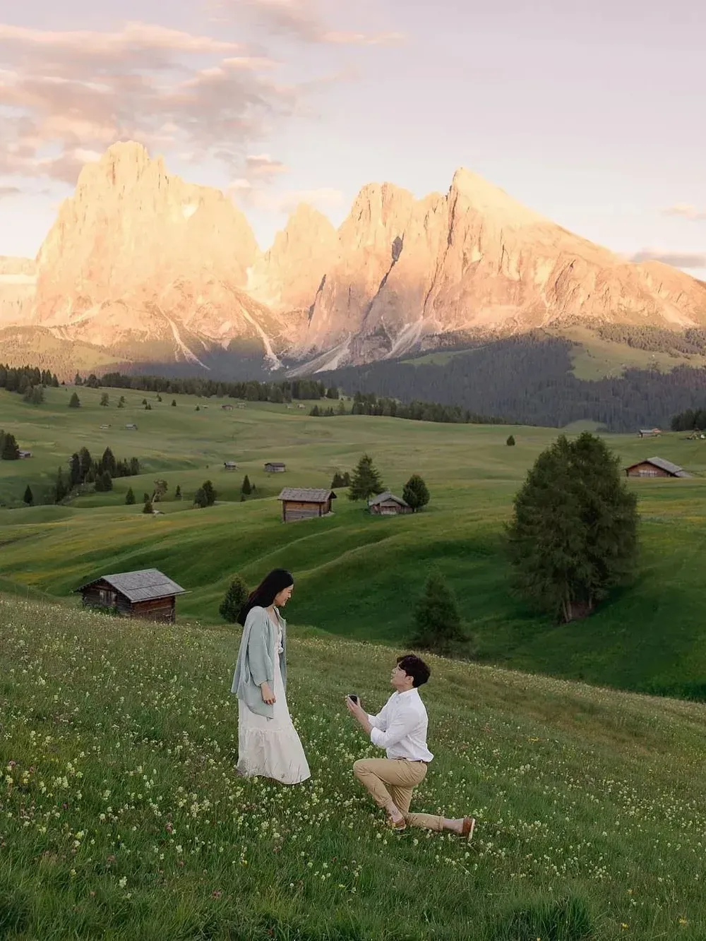 Couple in an Italian hillside meadow; one person kneels, proposing to the standing partner, with mountains and small huts on the horizon at sunset.