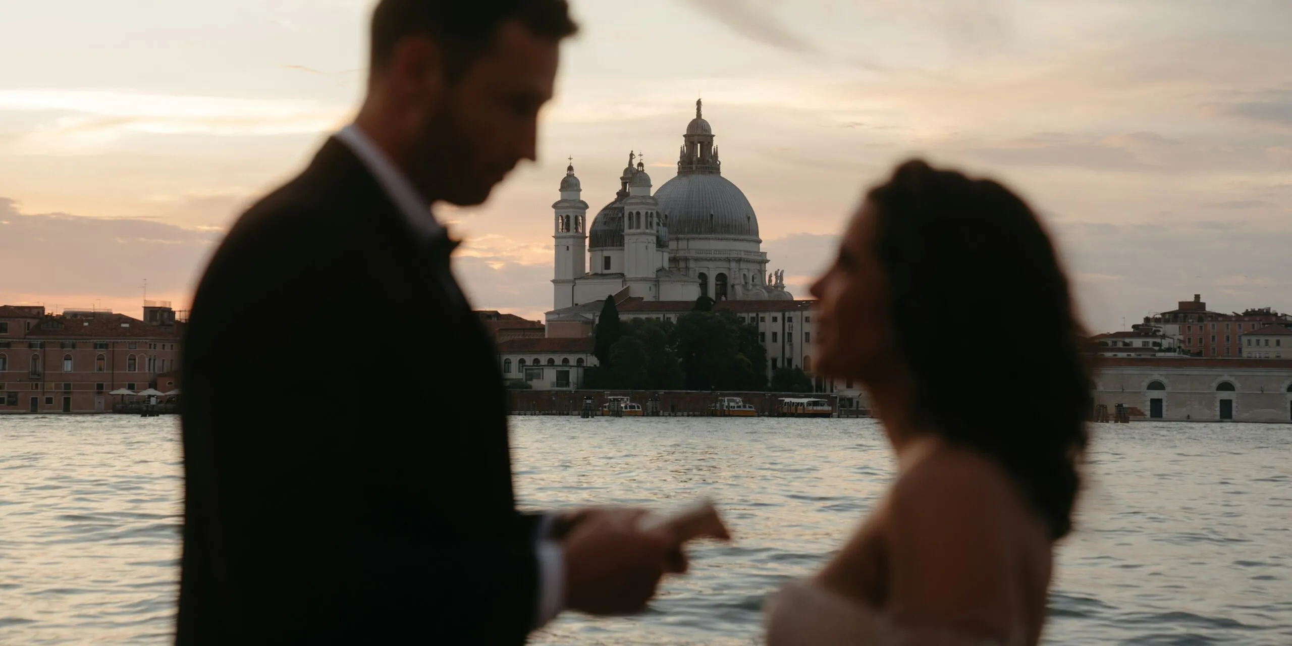 Blurred couple in formal attire by a sunset waterfront, with Venice's domed church and historic buildings across the water.