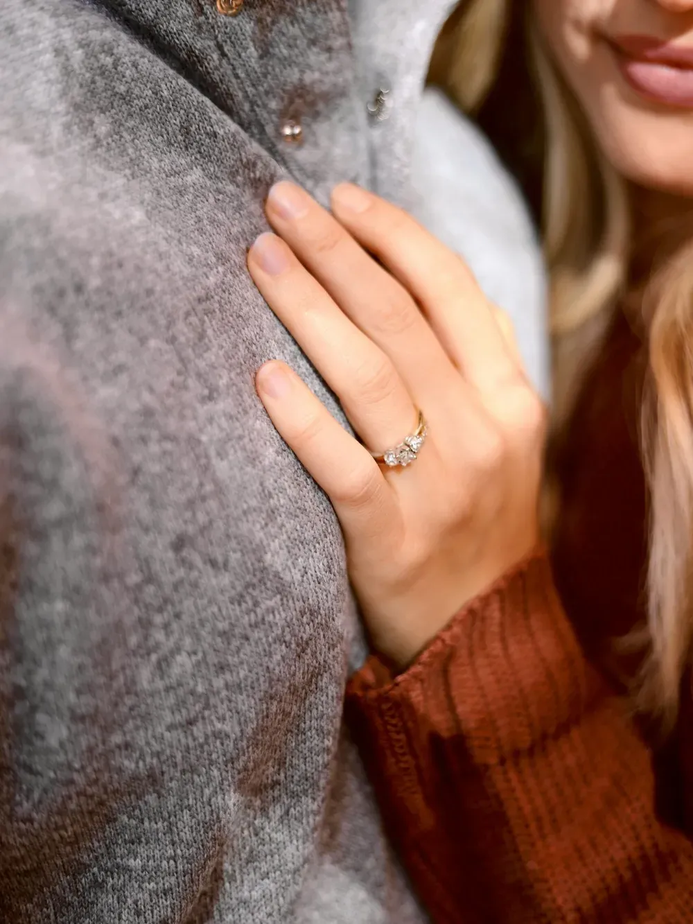 Close-up of a hand with a diamond ring resting on a gray textured coat; another person with blonde hair smiles nearby.