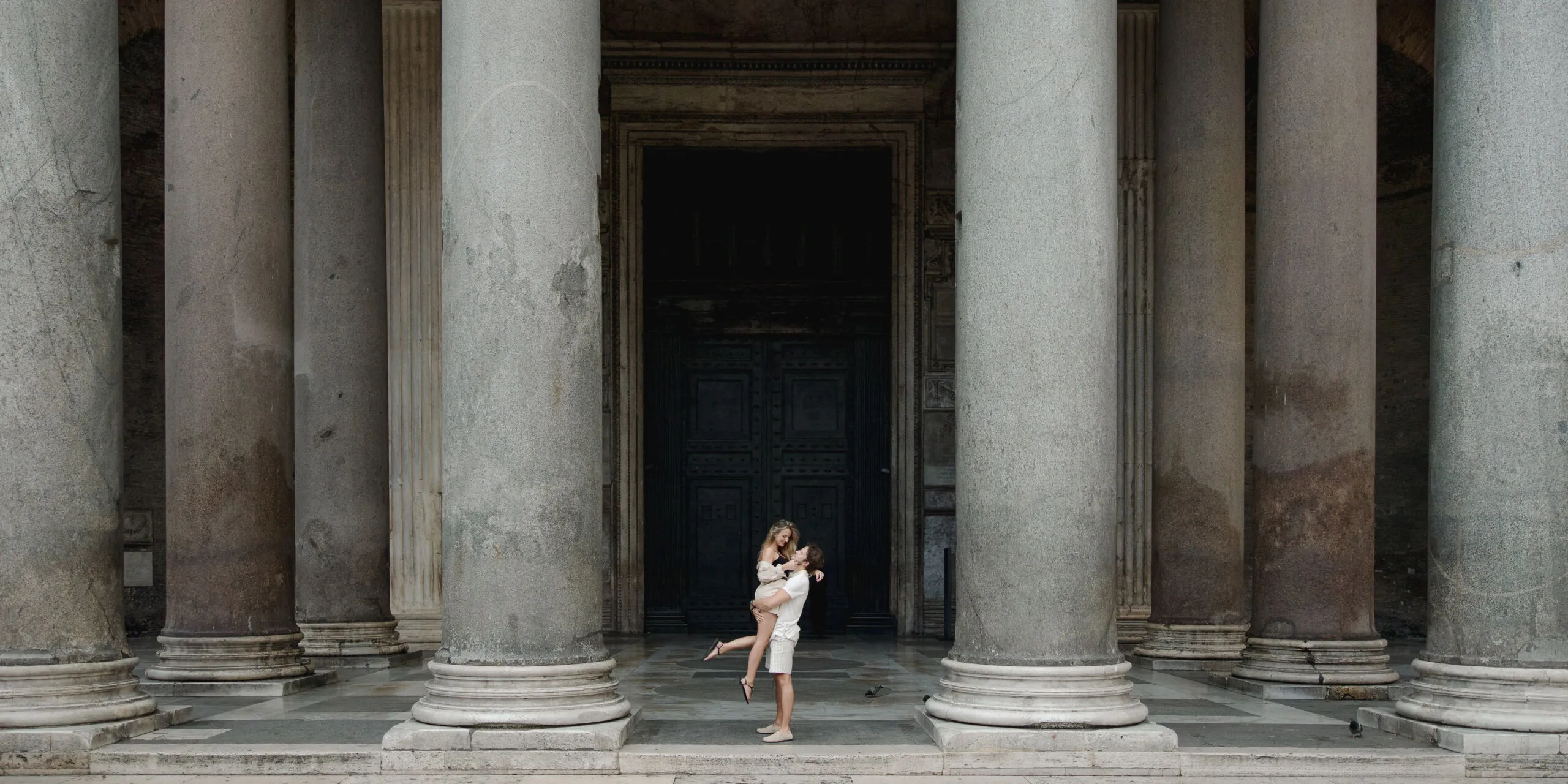 Two people embrace between tall stone columns and a dark doorway on a marble floor, in a classical Italian architectural setting.