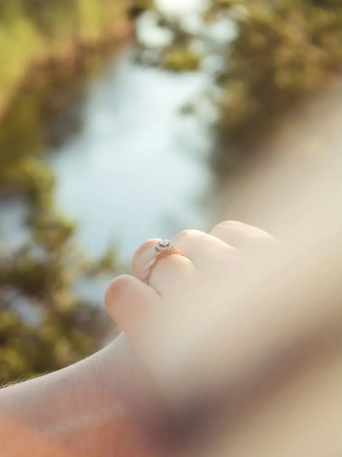 Close-up of a toe wearing a diamond engagement ring, with a soft-focused outdoor background of water and greenery.