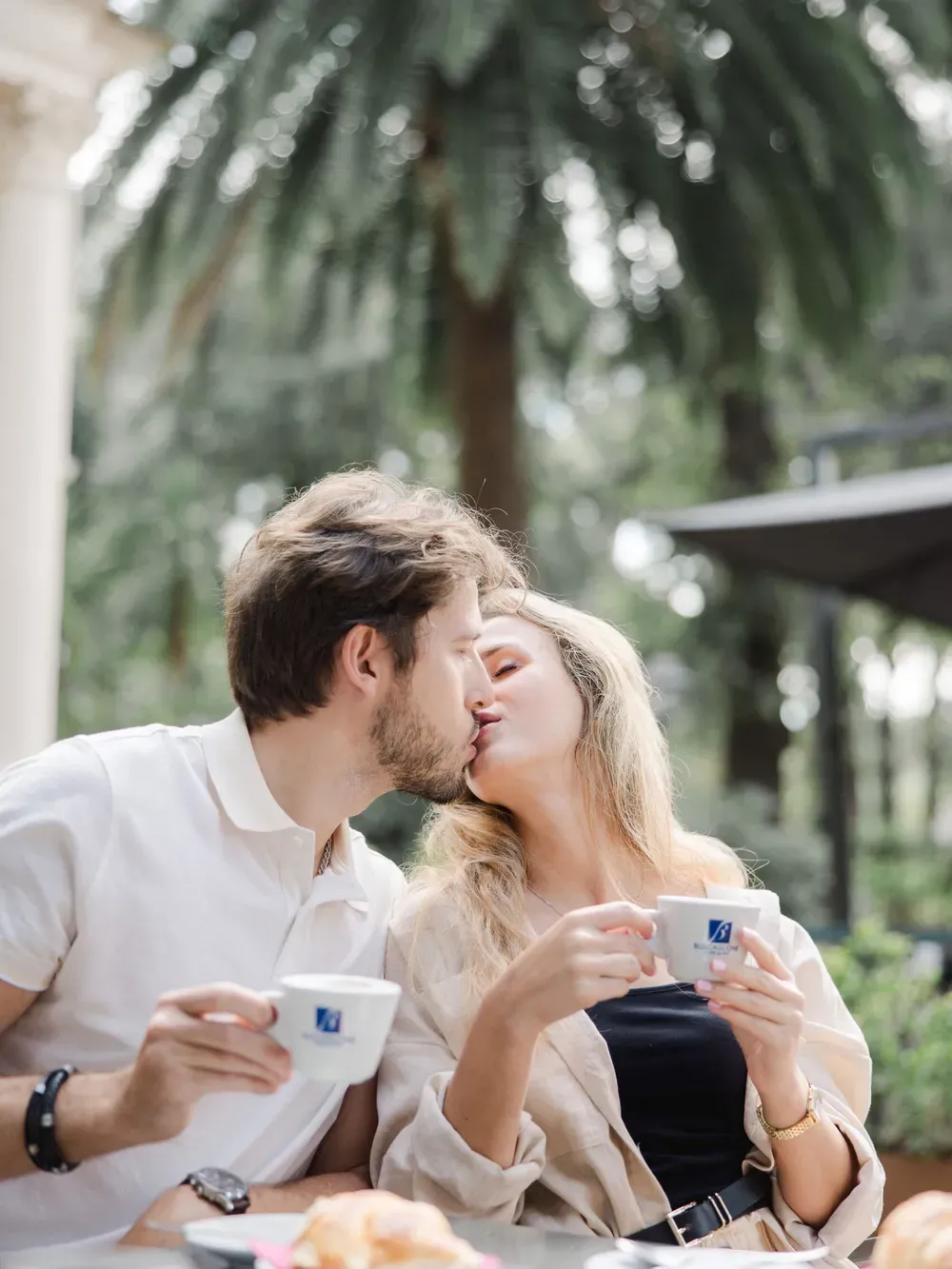 Two people kiss over coffee at an outdoor Italian cafe, with pastries on the table and lush greenery in the background.