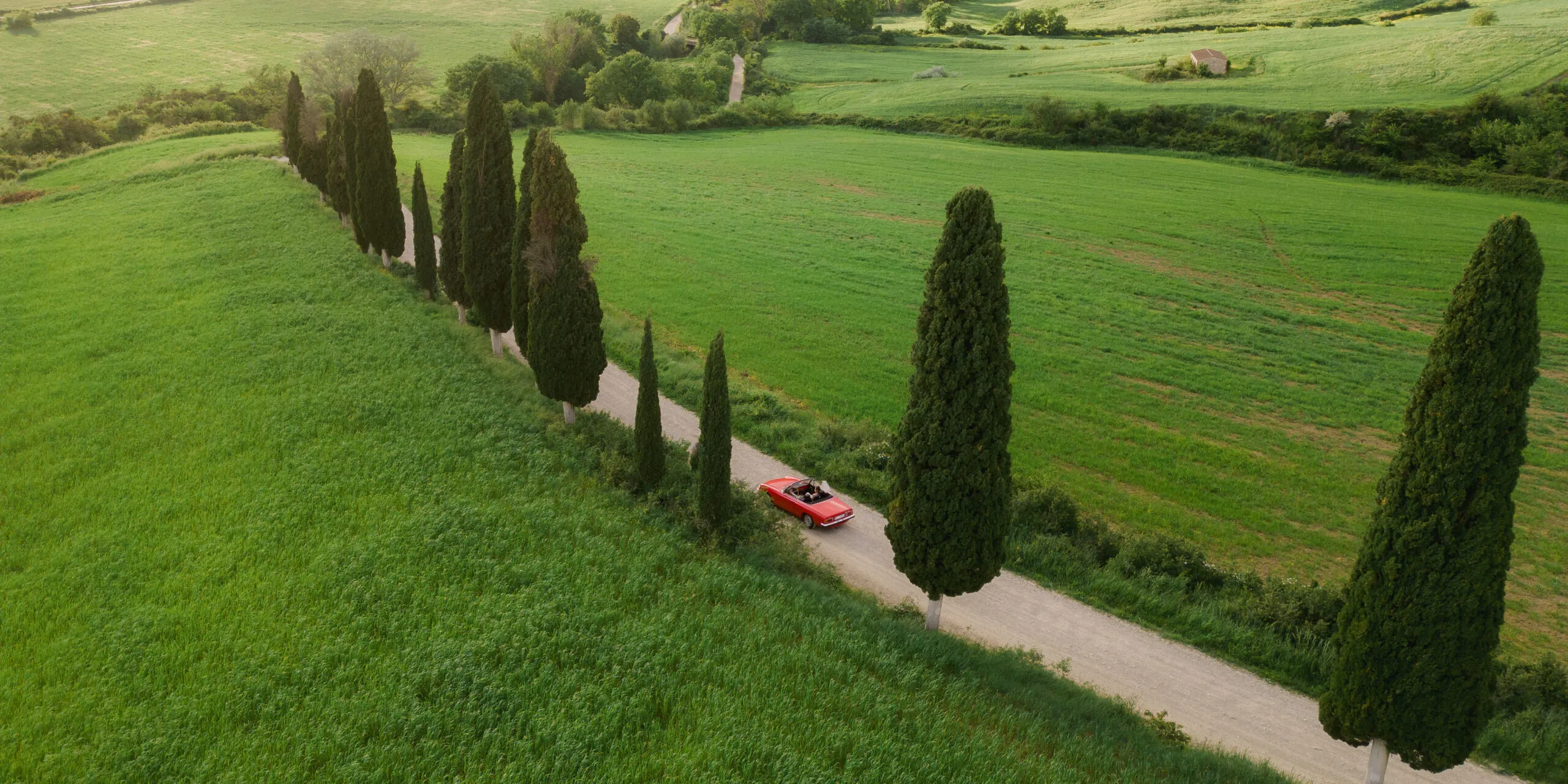 Red convertible on a narrow gravel road lined with tall cypress trees, winding through lush Tuscan hills—romantic Italian countryside.