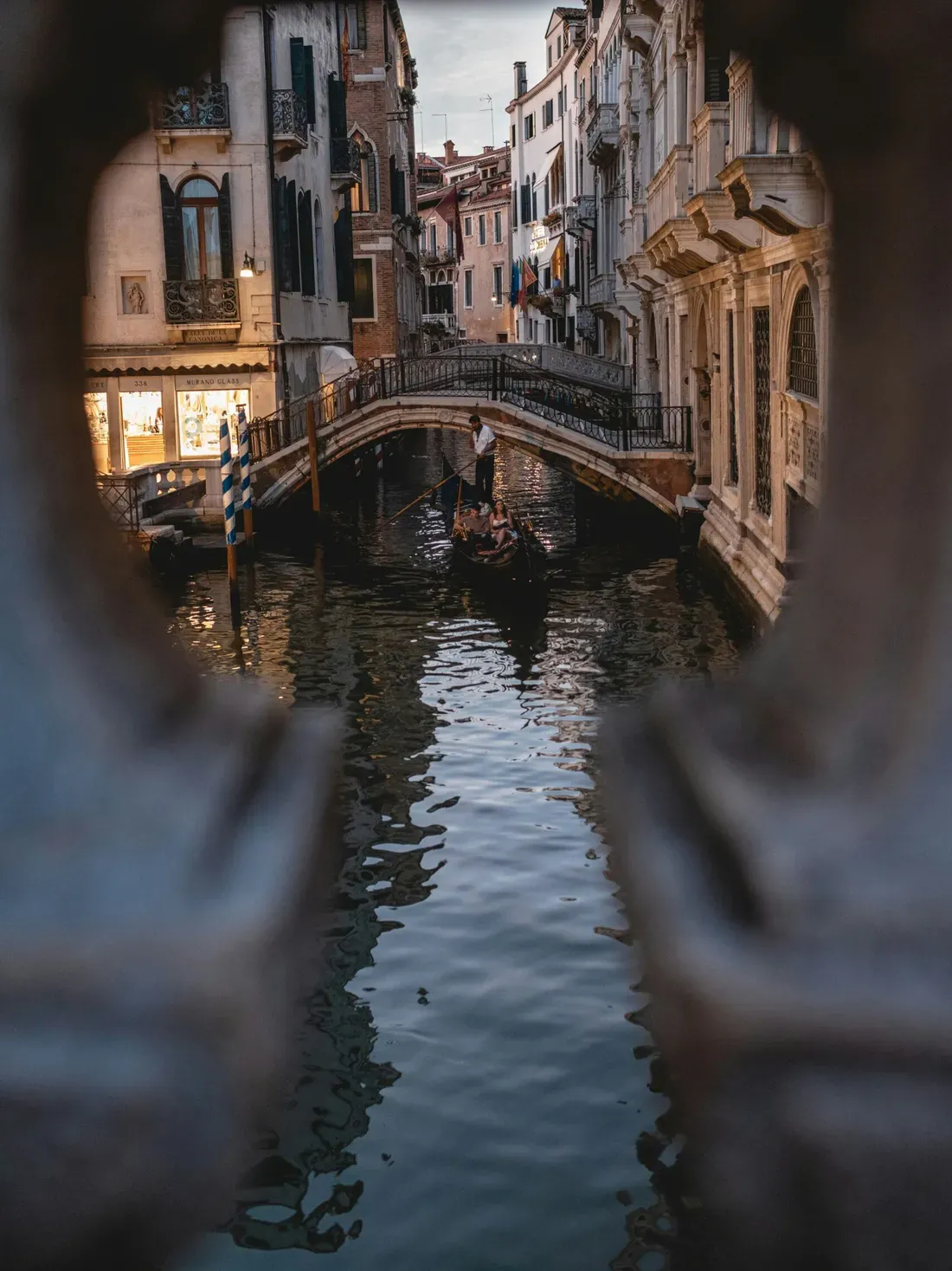 View through a stone arch of a romantic Venetian canal: a gondola with passengers beneath a bridge, lined by historic buildings at dusk.