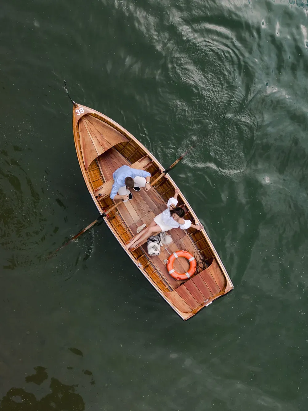 Top-down view of a small wooden rowboat with two people aboard, a bright orange life ring on the deck, drifting over dark green water.