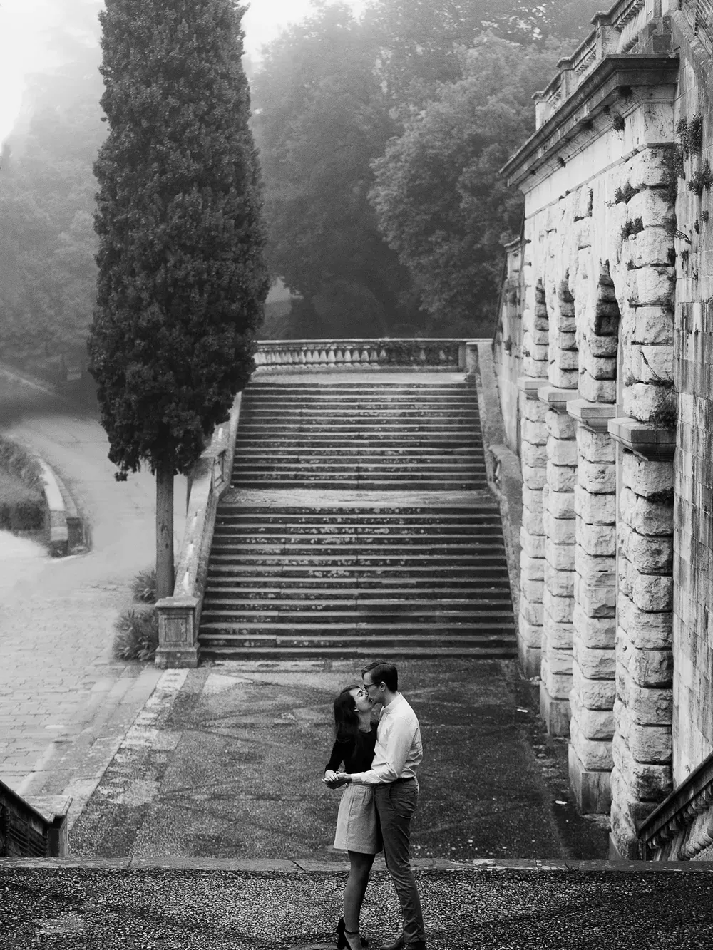 Black-and-white image of a couple embracing and kissing on a stone staircase beside a historic stone building, with tall trees and a misty background.