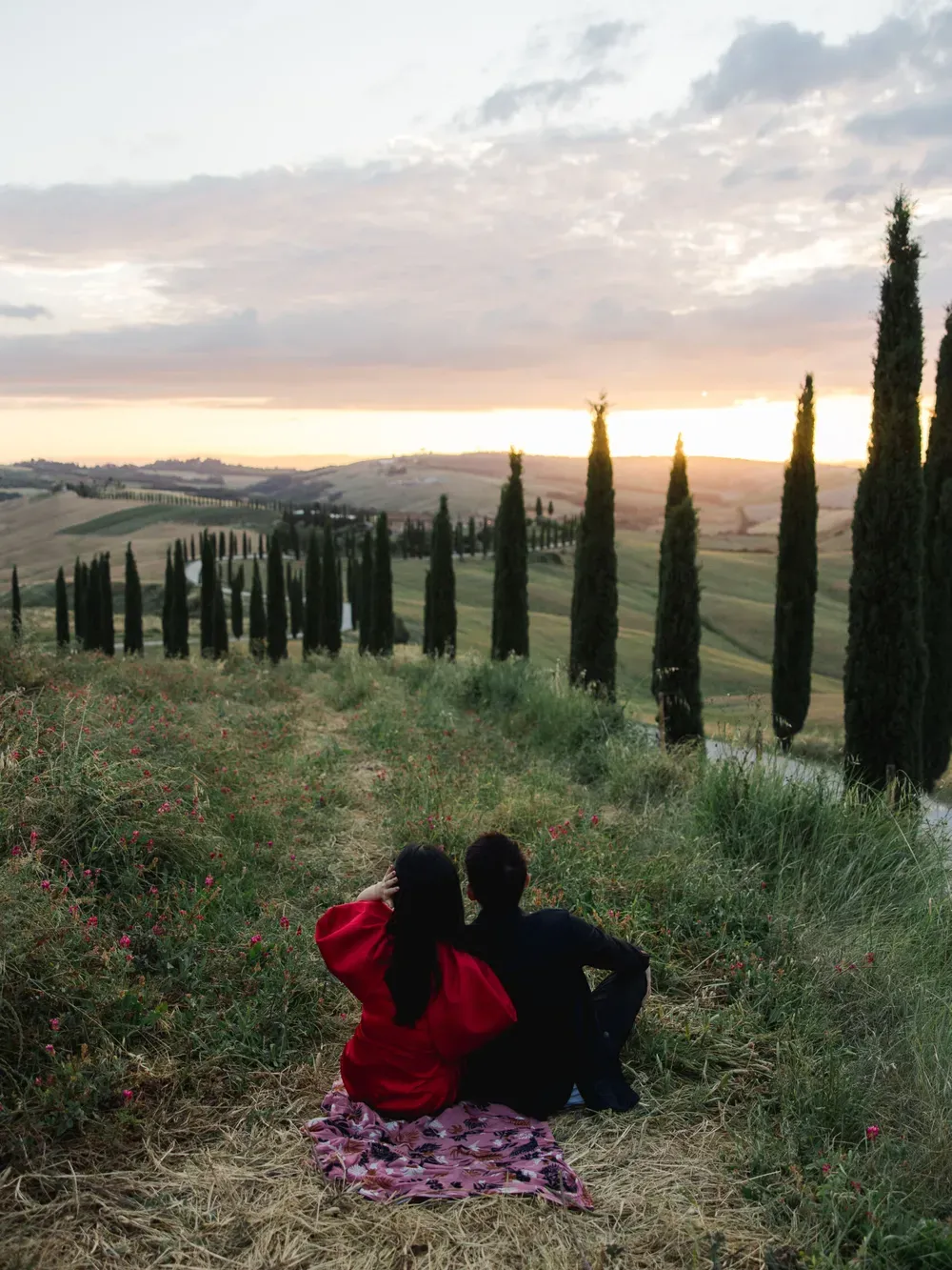 Couple sitting on a pink patterned blanket in a wildflower field at sunset, overlooking a Tuscan hillside with tall cypress trees.