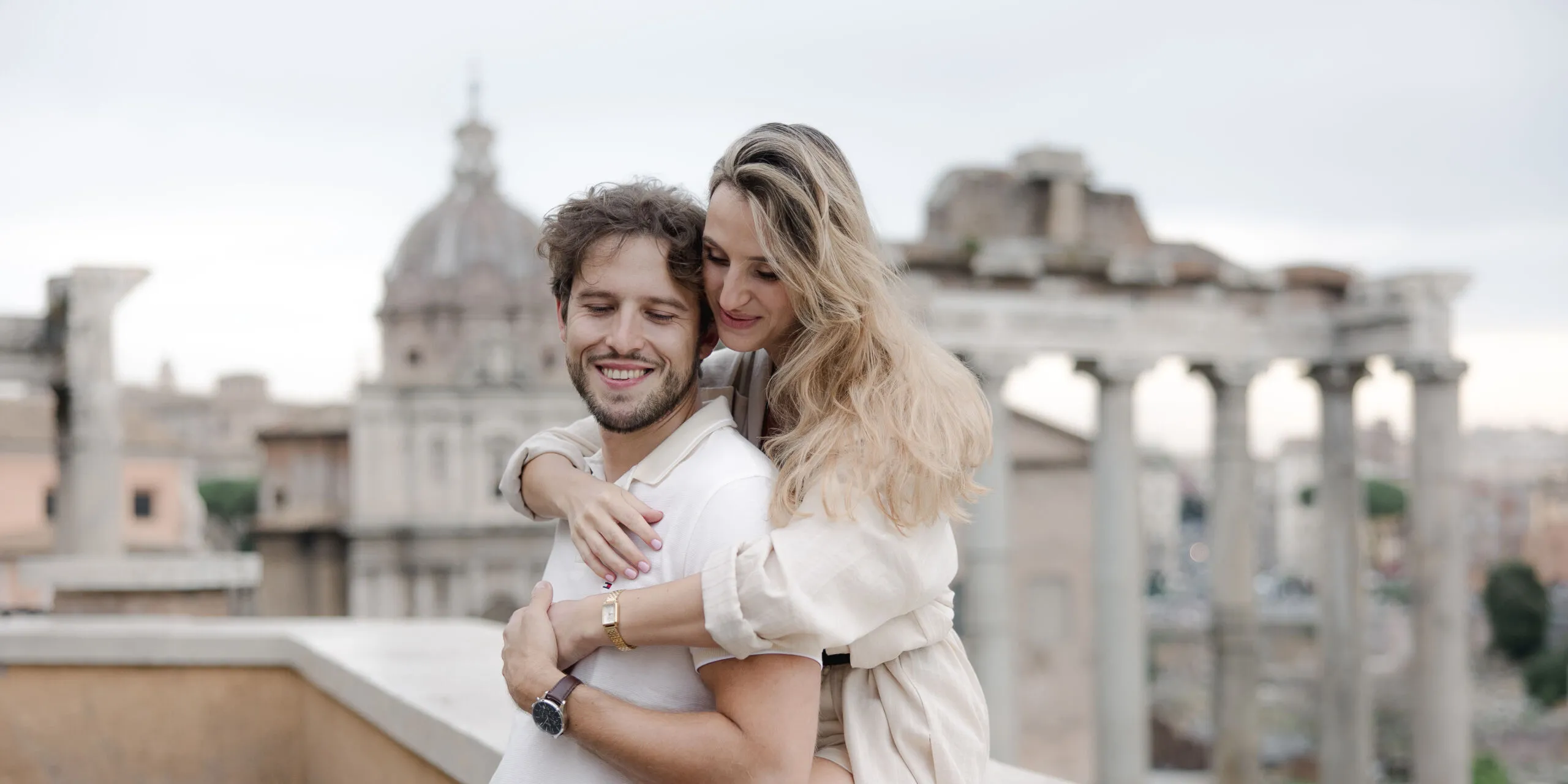 A couple hugs on a terrace in Italy, with ancient ruins and a domed building in the background; a straw hat sits on the ledge.
