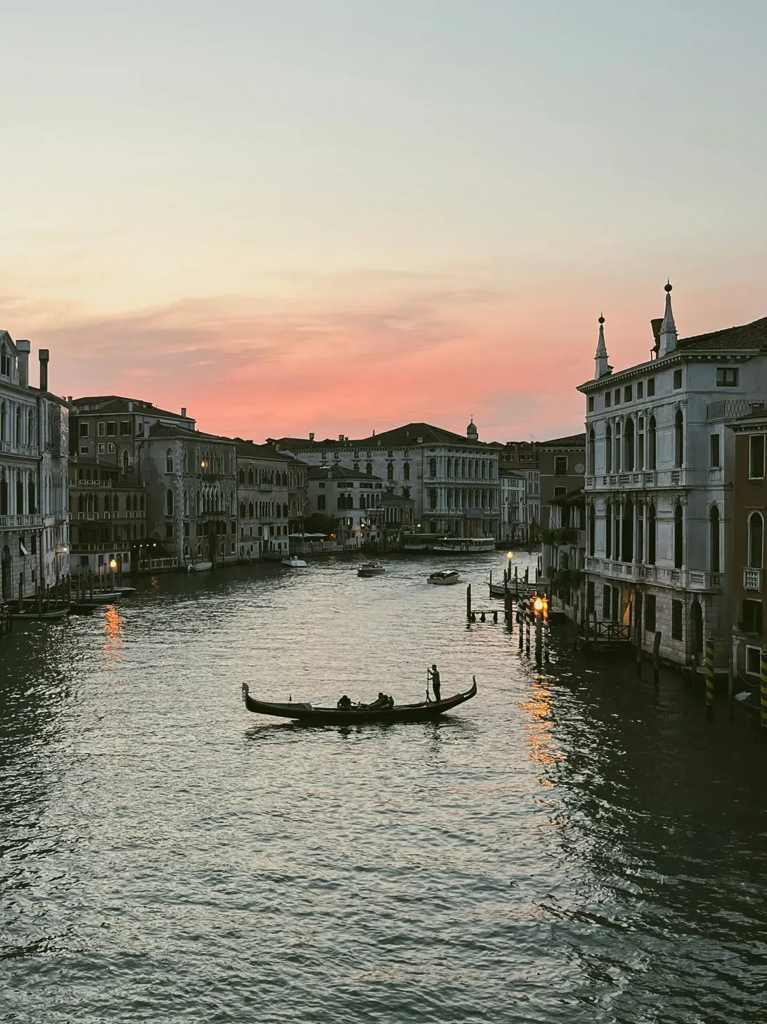 Romantic Venice canal at sunset in Italy, with a gondola carrying passengers, a gondolier steering, and historic buildings along glowing water.