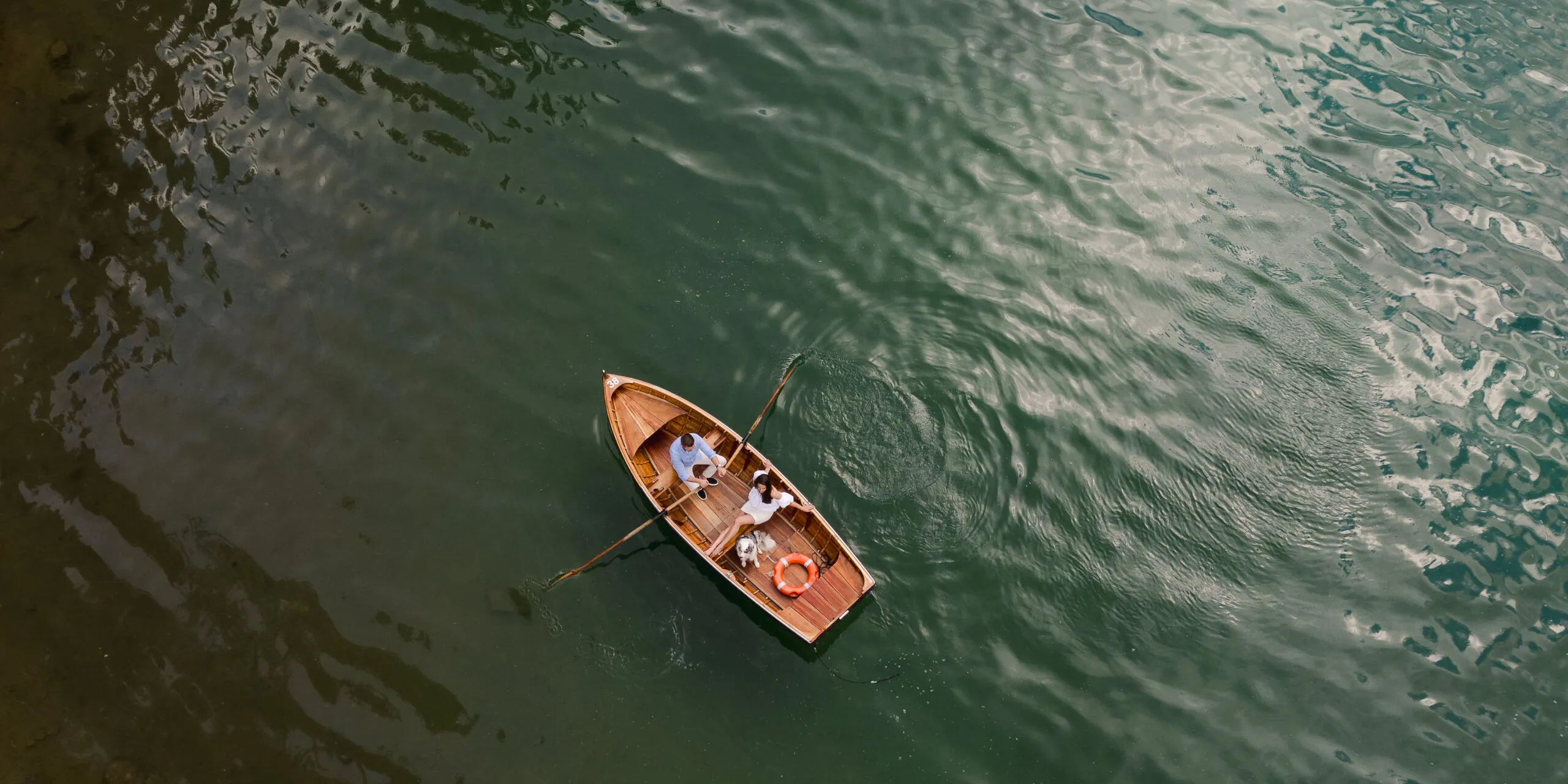 Aerial view of a small wooden rowboat with two people and an orange lifebuoy, afloat on green water with gentle ripples.