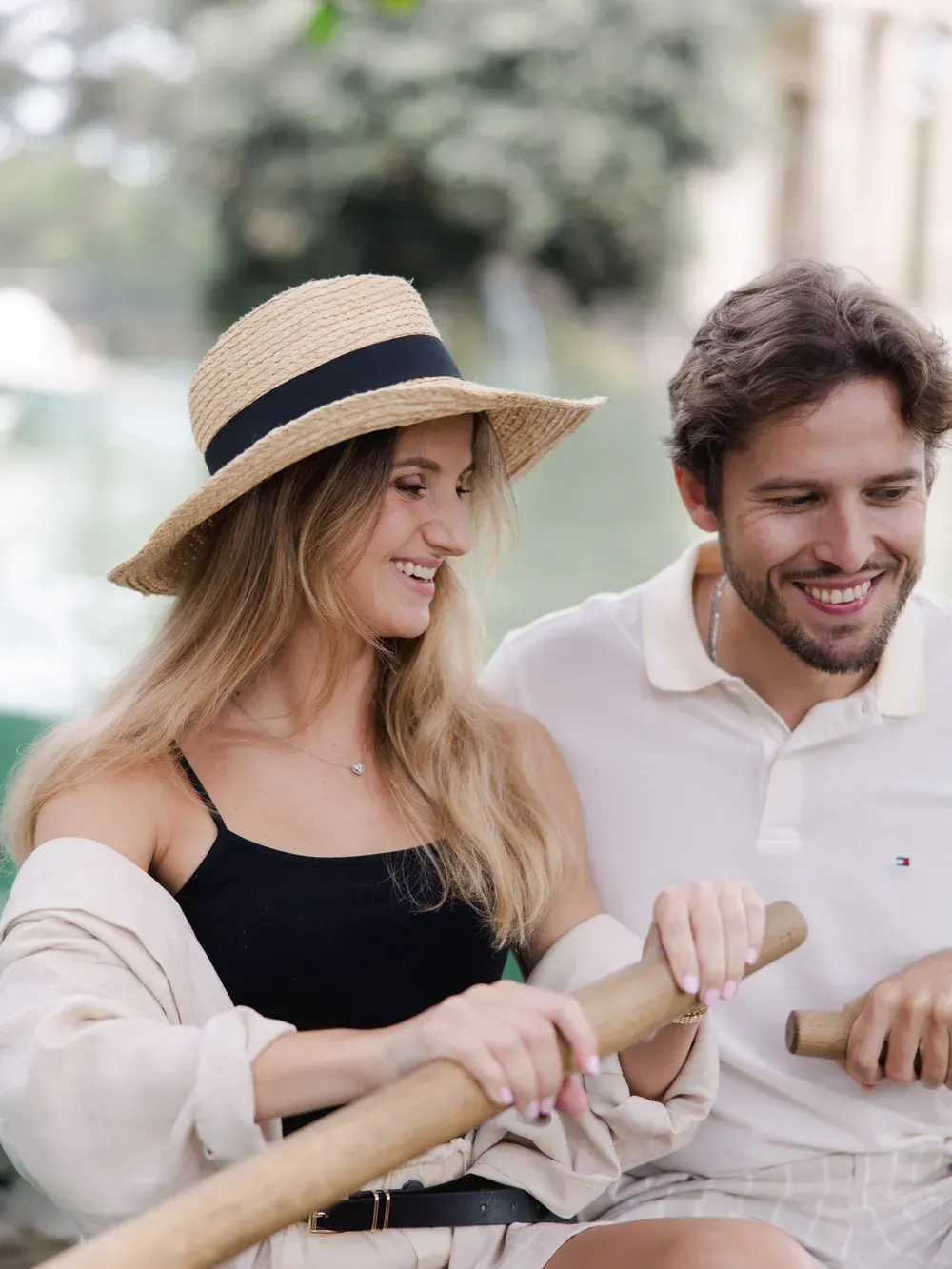 Two people smiling while rowing a boat on a sunny day in Italy; one wears a straw hat and black top, the other a light polo, with greenery behind.