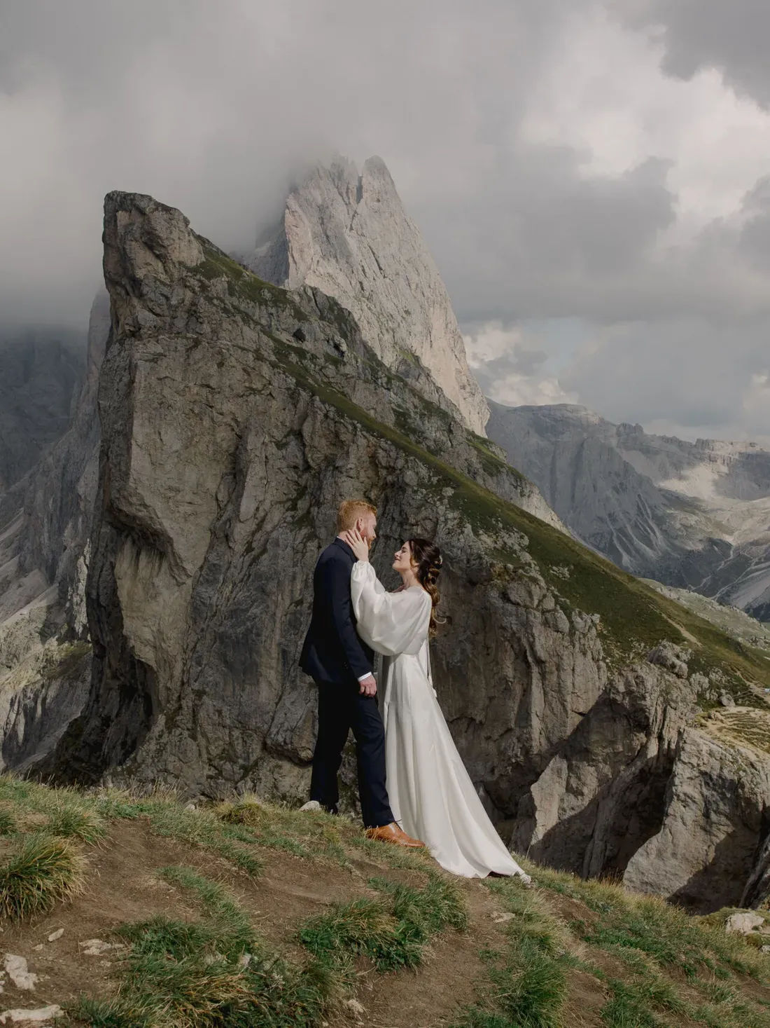 A couple in wedding attire embraces on a grassy alpine ledge, with dramatic rocky peaks and a cloudy sky in the background.