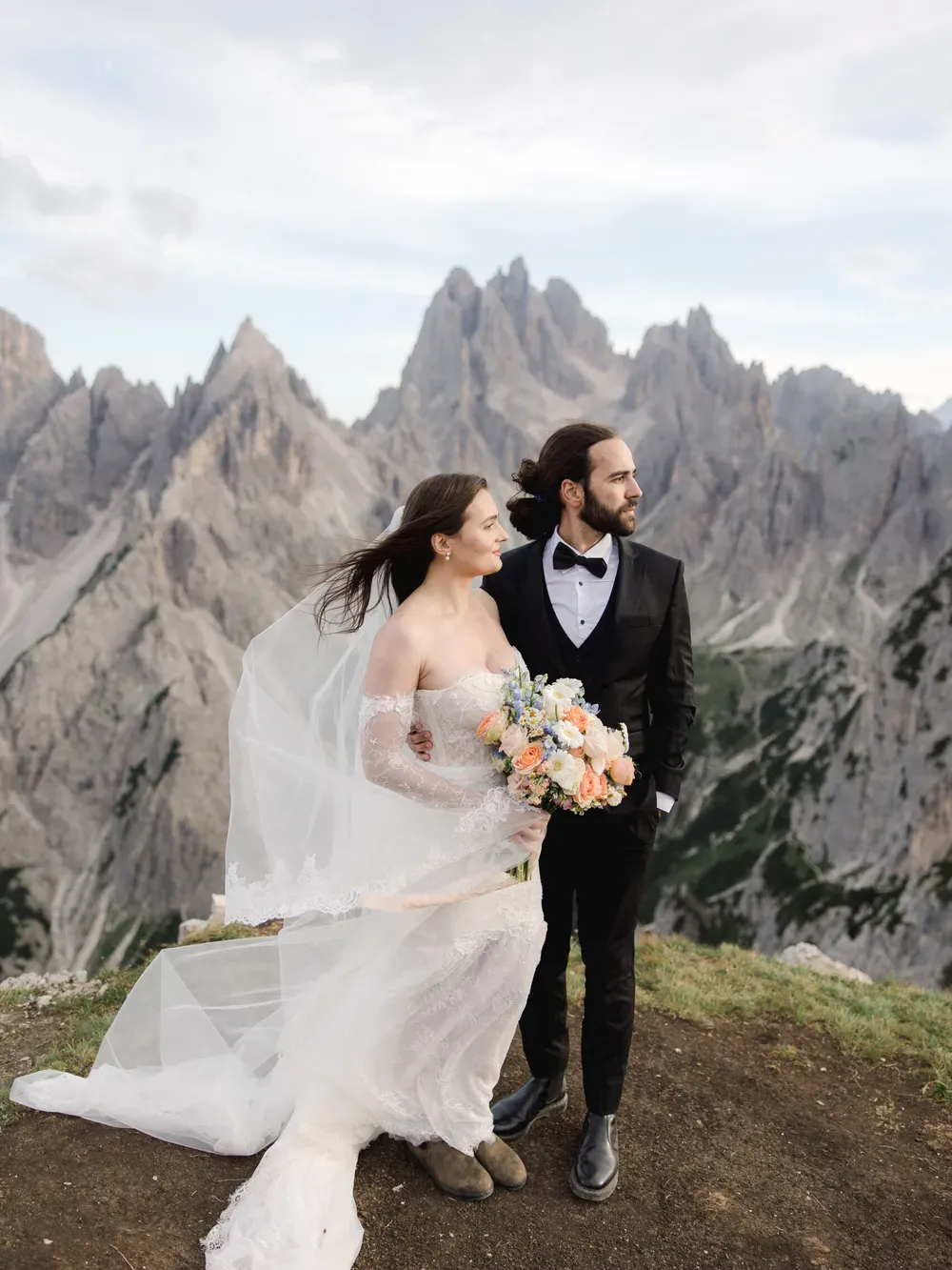 Two people in wedding attire stand on a rocky cliff with jagged mountains in the background; one holds a pastel bouquet as a veil flows.