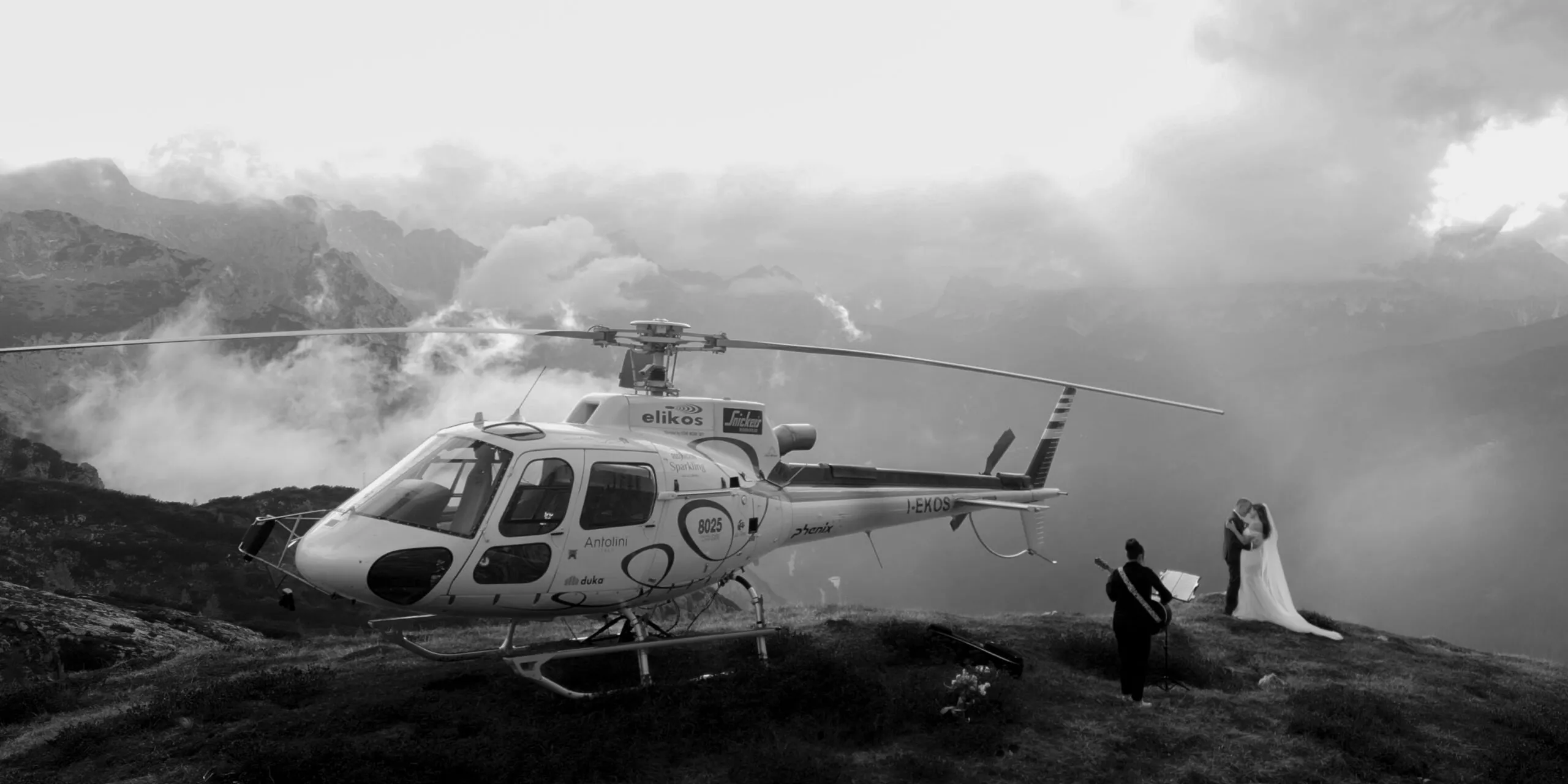 A helicopter sits on a grassy mountain ledge as a couple in wedding attire pose with a photographer against a misty, rugged alpine landscape.