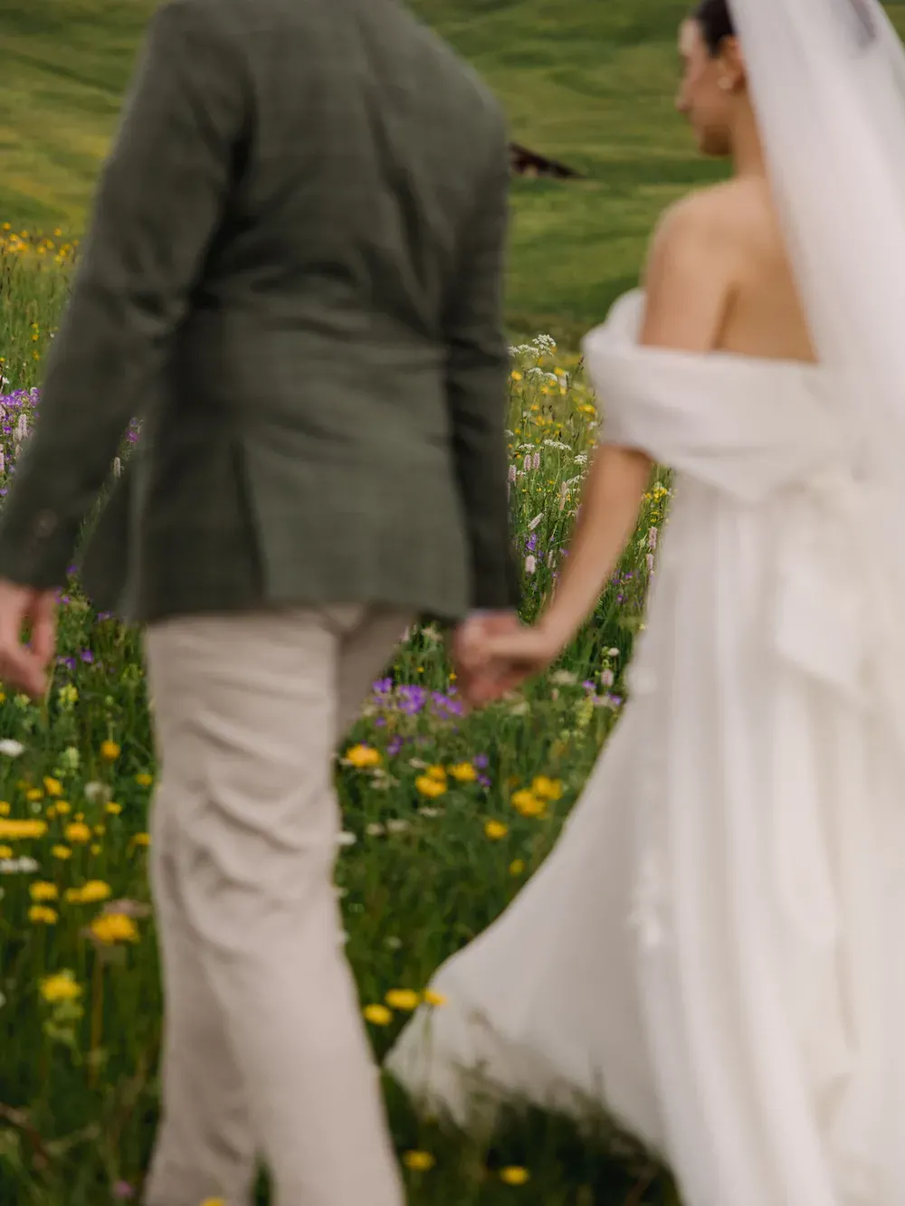 Couple walking hand-in-hand in an Italian meadow; one in a wedding dress with veil, the other in a green blazer and light pants, seen from behind.