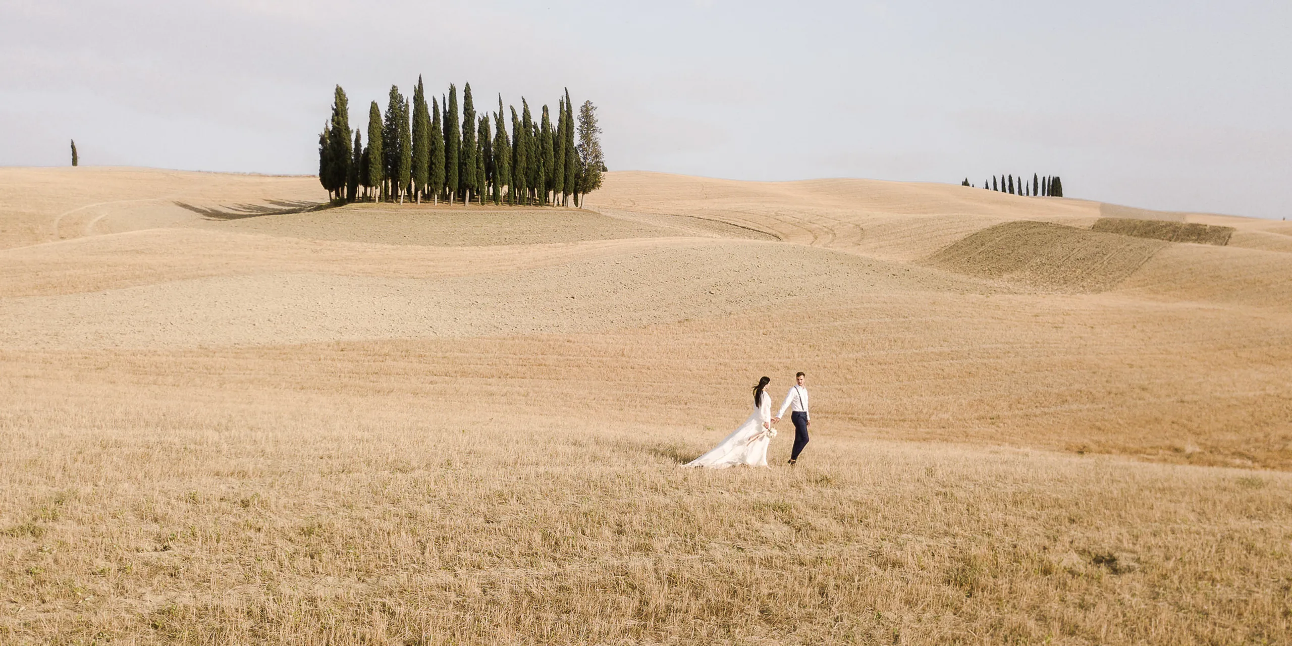 Two people in wedding attire walk across a golden Italian countryside field, with a line of tall cypress trees on a hill under a pale blue sky.