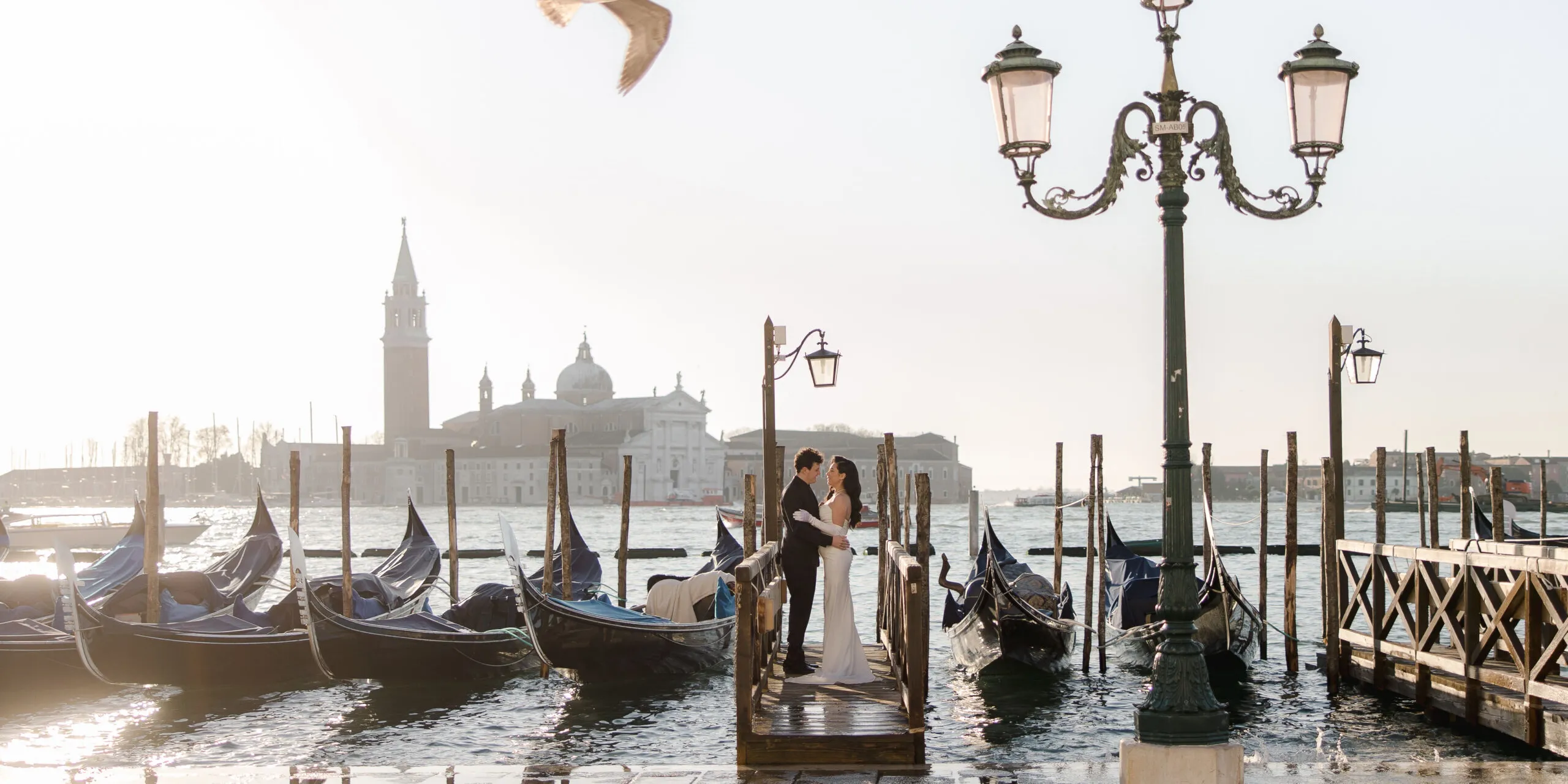Romantic couple in wedding attire on a wooden dock among gondolas in Venice, with a vintage lamp post and distant domed church in the background.