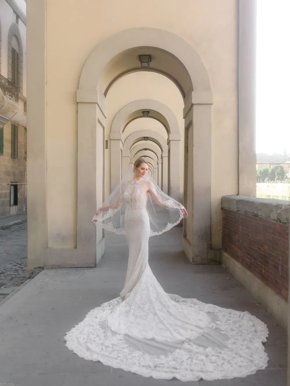 Person in a white lace wedding gown with a long train and veil stands in a sunlit colonnaded archway along an Italian walkway.