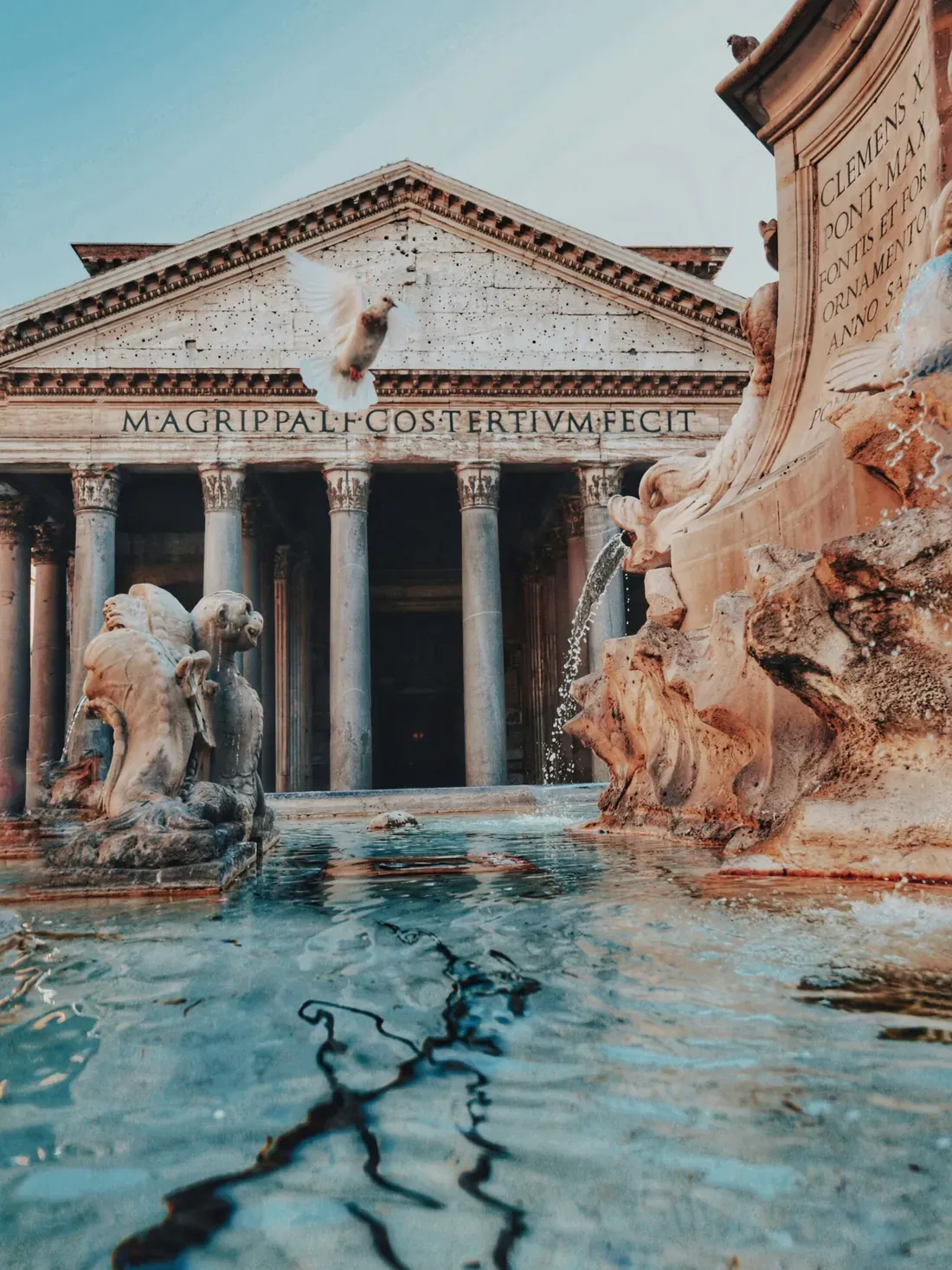 Rome-style temple facade with tall columns and a triangular pediment, behind a large fountain; a bird flies over the scene.