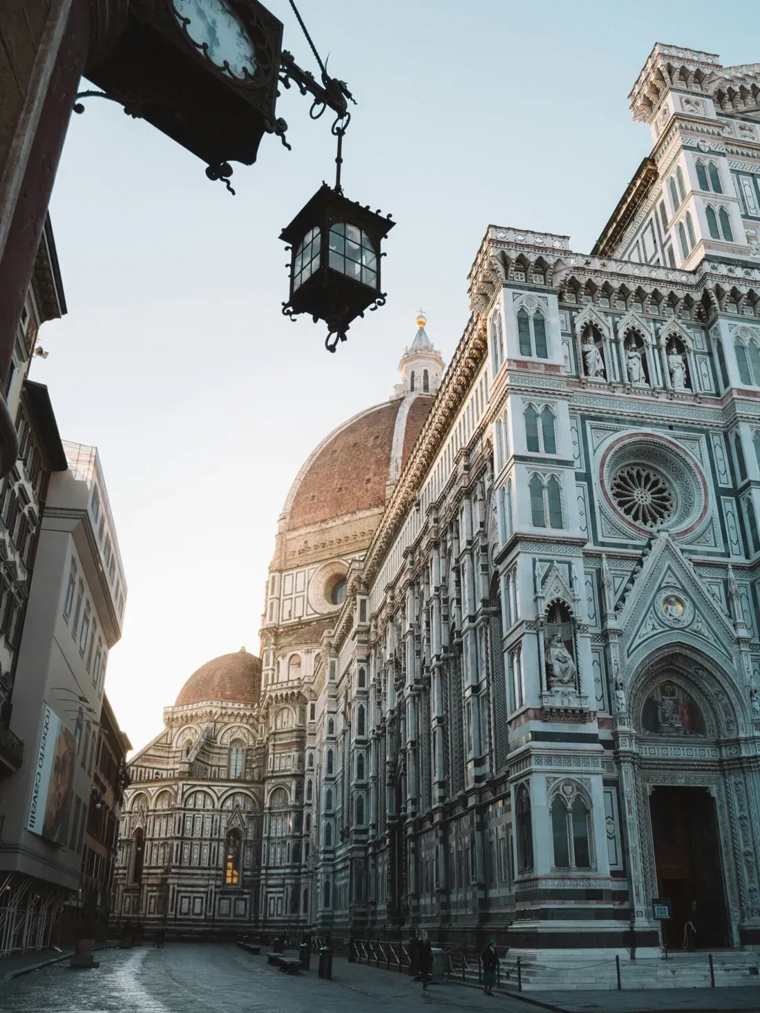 Sunlit Florence street scene with the cathedral’s ornate marble facade and red dome, and a hanging lantern in the foreground.