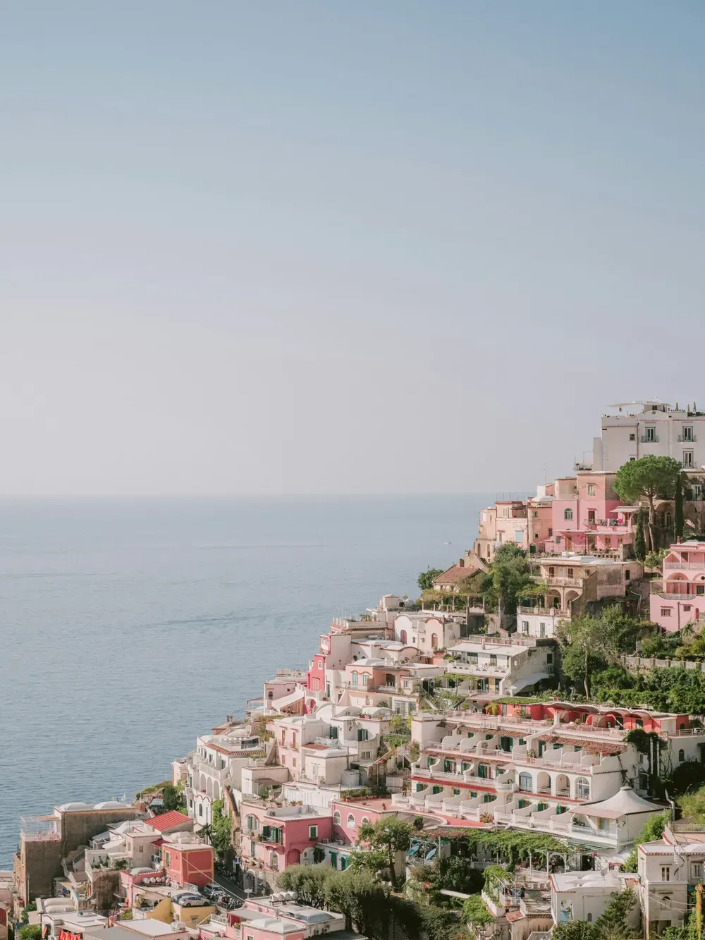 Pastel-colored Italian hillside town along the coast, with white, pink, and peach buildings stacked on terraces above a calm blue sea.