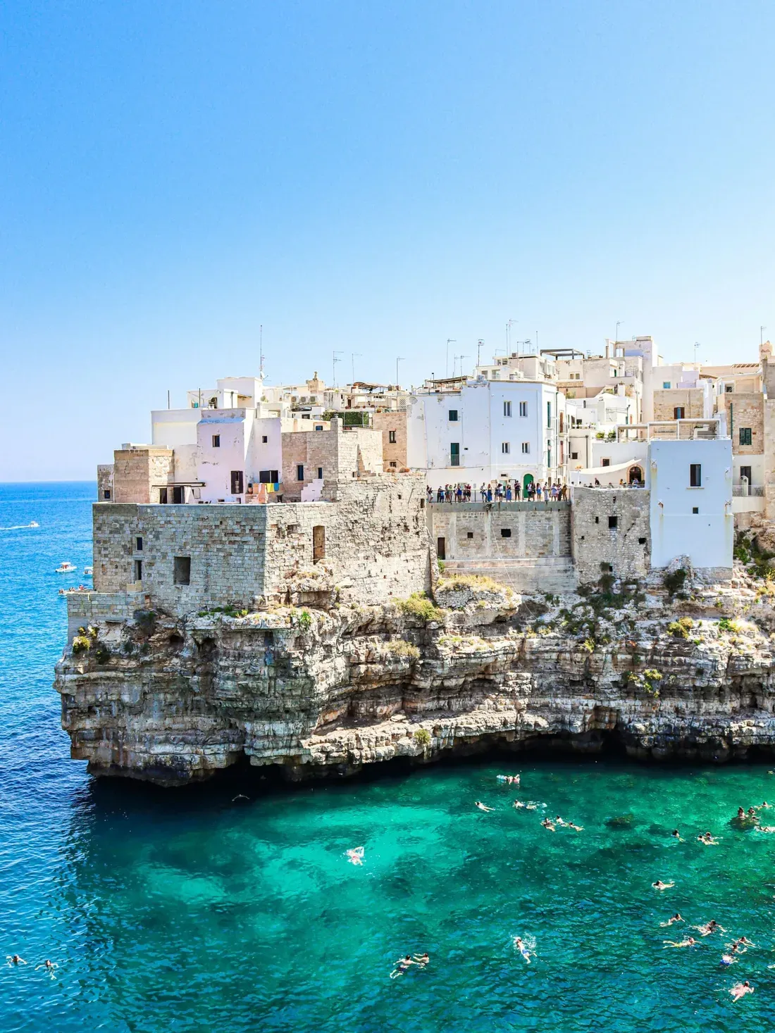 Whitewashed buildings cling to a rocky Italian cliff above turquoise sea, with a stone fortress and people along the edge under a clear blue sky.