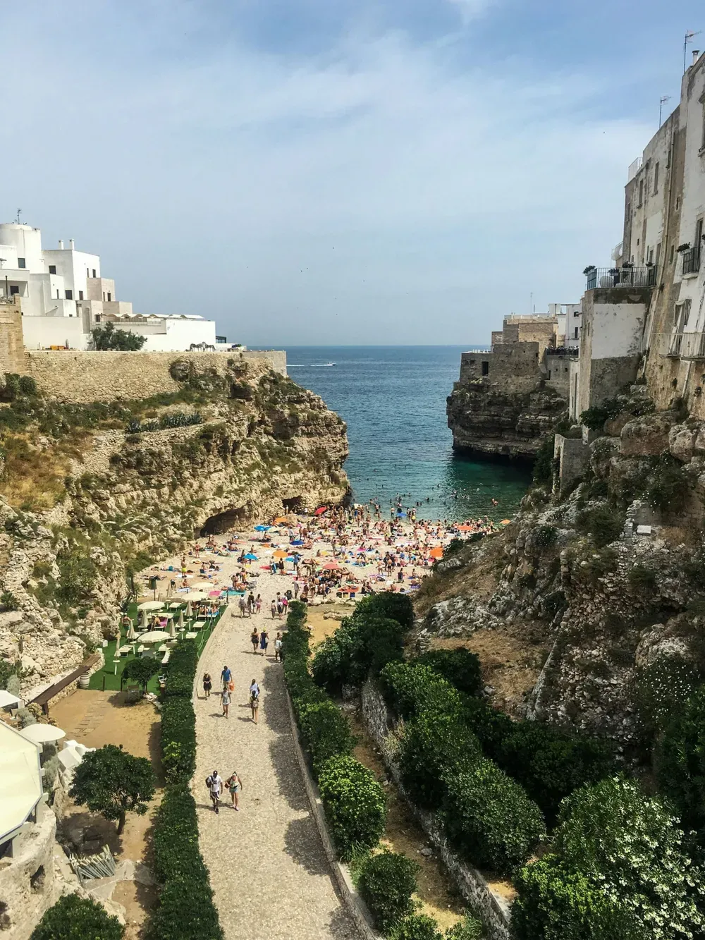 Cliffside Italian town with whitewashed buildings, a pebble beach crowded with sunbathers, and turquoise sea beside a cobblestone promenade.