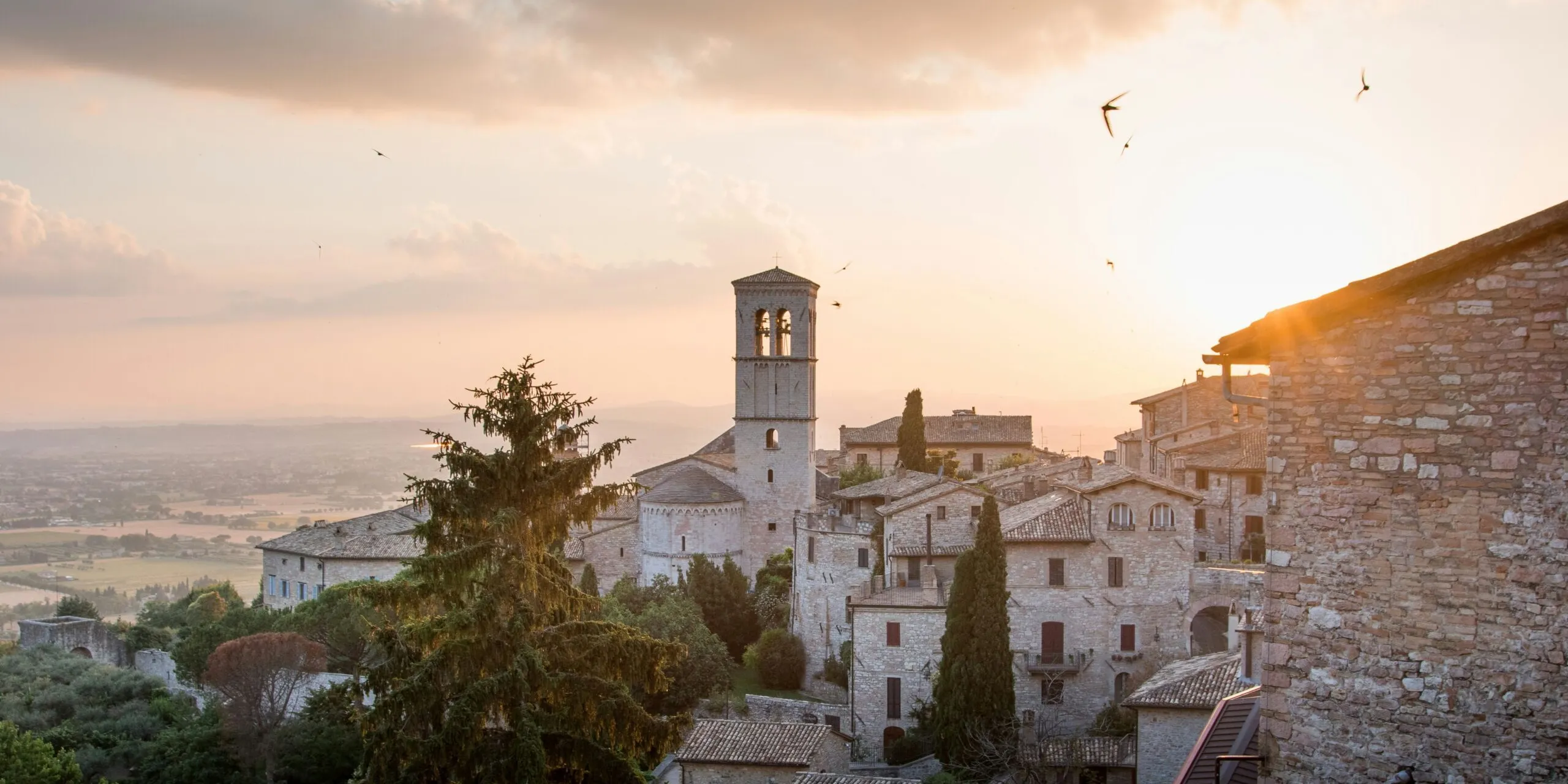 Sunset over a historic Italian hill town with stone buildings, a tall bell tower, terracotta roofs, and surrounding cypress trees.