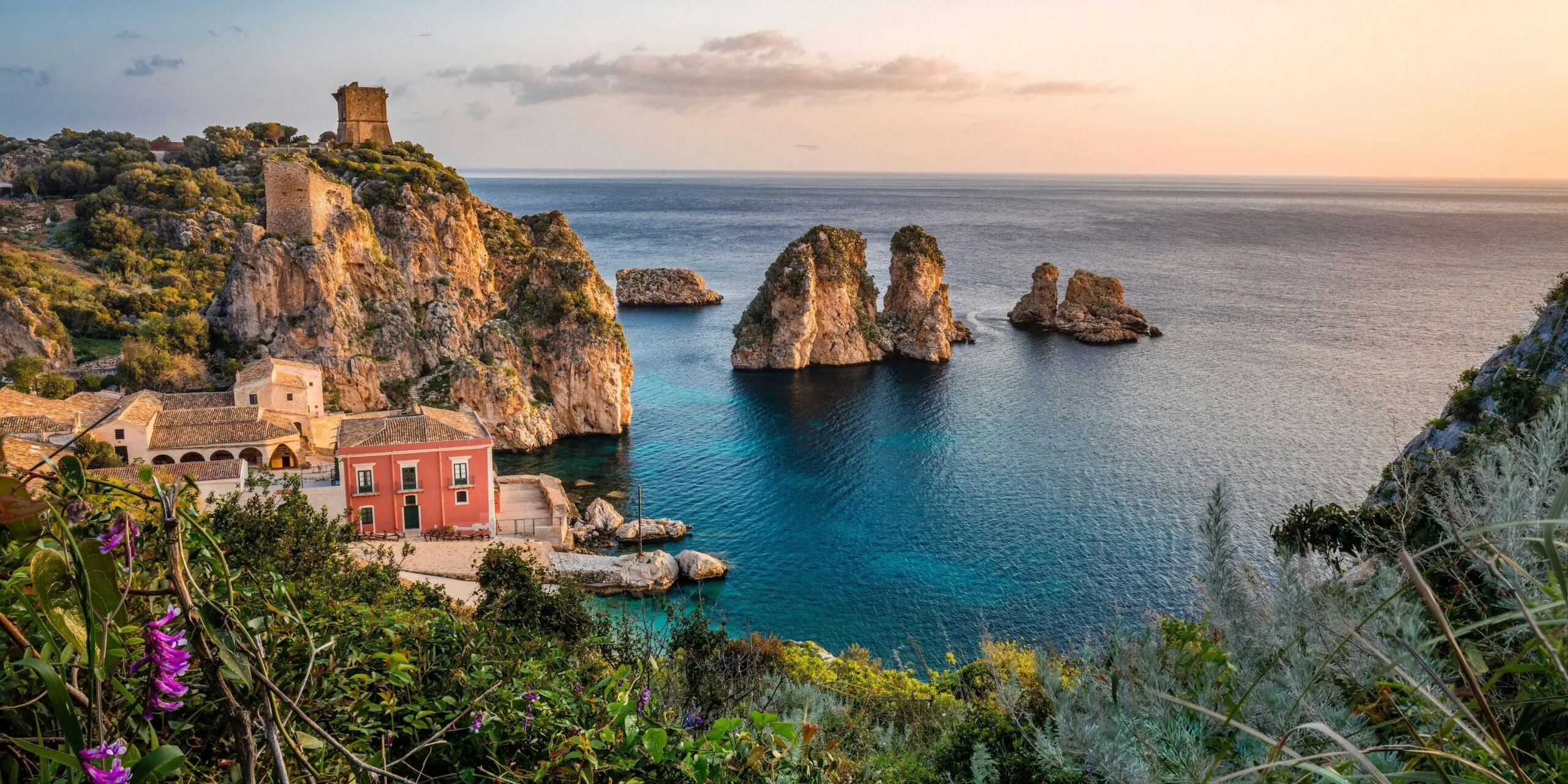 Sunset over an Italian Mediterranean coastline with rocky cliffs, turquoise water, a pink building by the shore, and lush foreground vegetation.