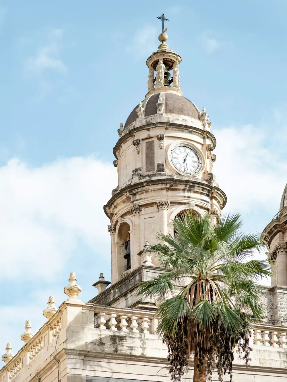 Italian clock tower with a domed top, cross, and intricate stonework, set against a blue sky, with a foreground palm tree.