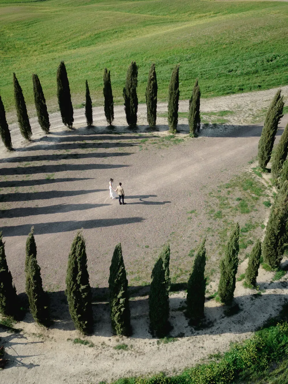 Aerial view of a circular row of tall cypress trees enclosing a gravel clearing, with two people holding hands at the center in the Italian countryside.