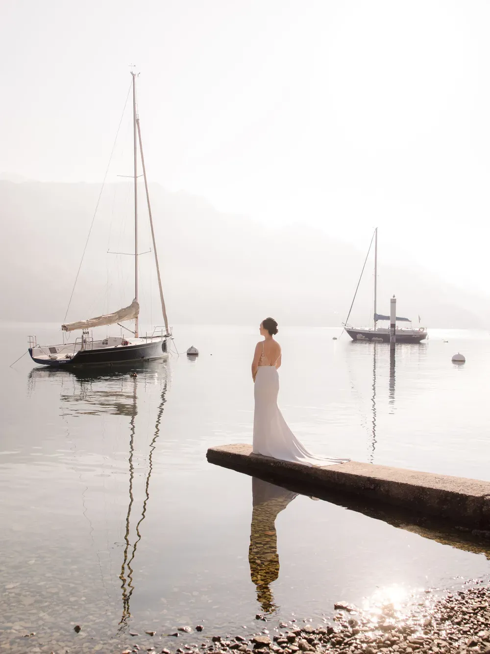 A person in a white gown stands on a dock by calm water in Italy, with sailboats and hazy mountains in the background.