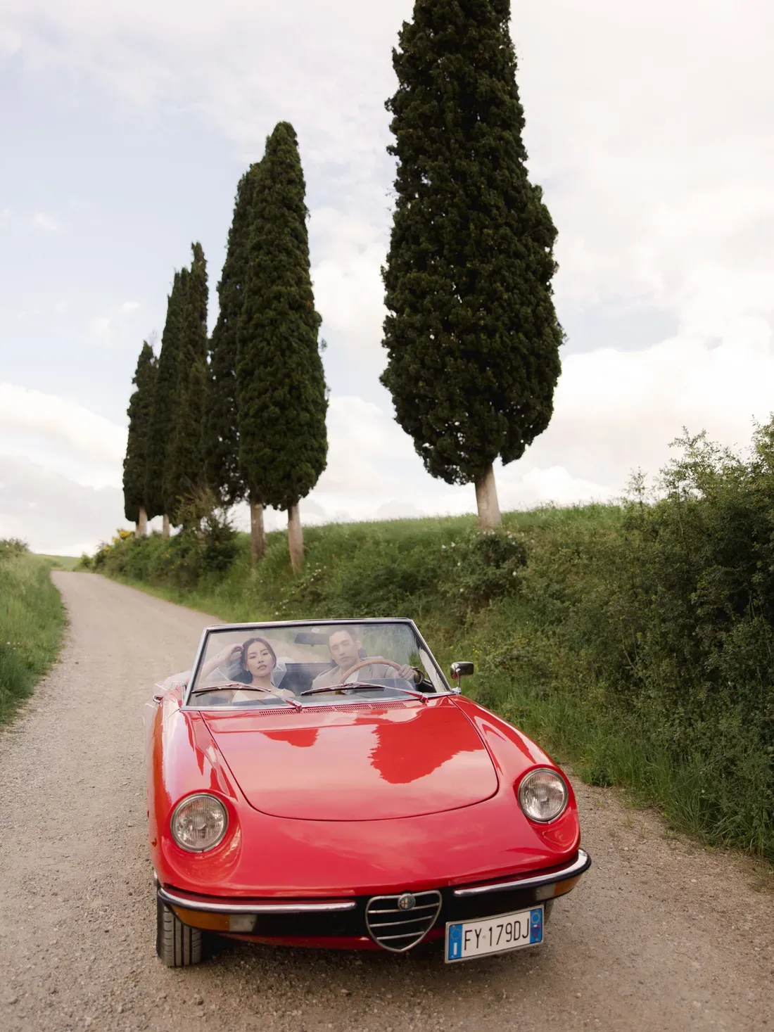 Red vintage convertible with two people driving a gravel country road, flanked by tall cypress trees in the Italian countryside.